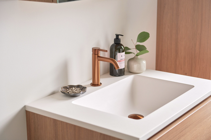 Salle de bain moderne avec vasque blanche et robinet coloré en bronze brossé, associée à un meuble en bois et des accessoires.