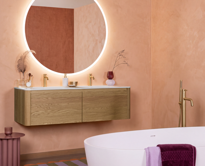 Badkamer in hippe kleuren met afgerond houten badkamermeubel en goudkleurig kraanwerk met een grote ronde spiegel.