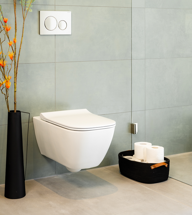 Salle de bain moderne avec WC blanc, carrelage mural vert, vase noir avec branches et panier de rouleaux de papier toilette.