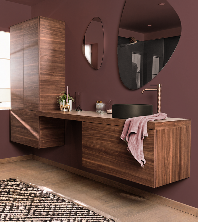 Tendance salle de bain avec meuble en noyer, vasque noire à poser, robinet en cuivre et miroirs organiques sur un mur bordeaux.