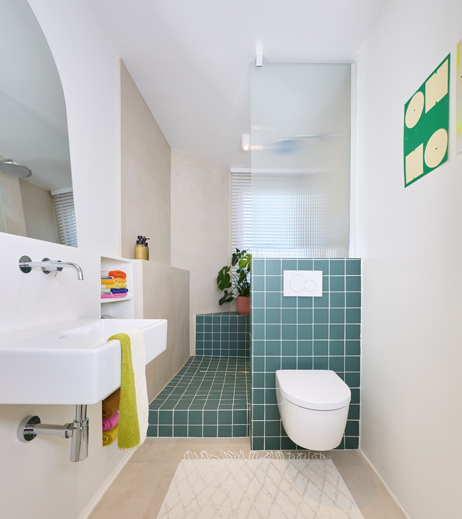 Salle de bain moderne verte avec WC suspendu, douche ouverte avec carreaux verts et lavabo blanc avec serviettes colorées.