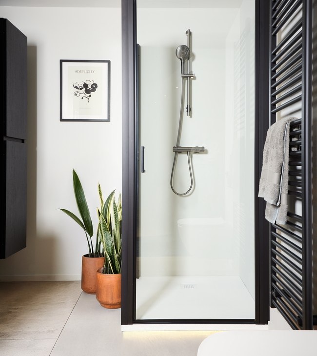 Douche moderne avec des accents noirs et un receveur de douche blanc en acrylique, entourée de détails élégants et de plantes d’intérieur.