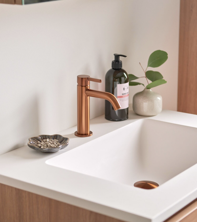 Salle de bain moderne avec vasque blanche et robinet coloré en bronze brossé, associée à un meuble en bois et des accessoires.