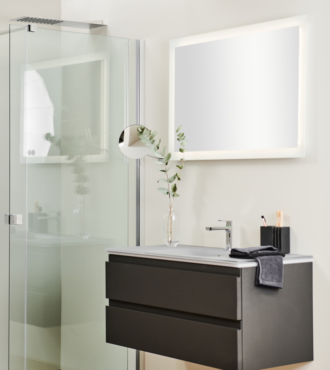 Meuble de salle de bain de style moderne avec meuble suspendu gris foncé, miroir éclairé et paroi de douche en verre sur un sol en bois clair.