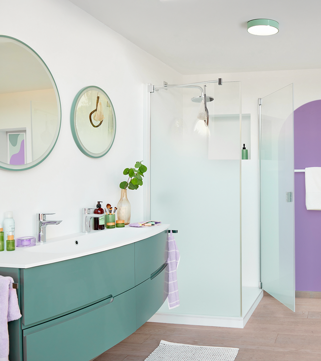 Salle de bain moderne avec cabine de douche en verre dépoli, meuble de bain vert et miroirs ronds dans des tons pastel frais.
