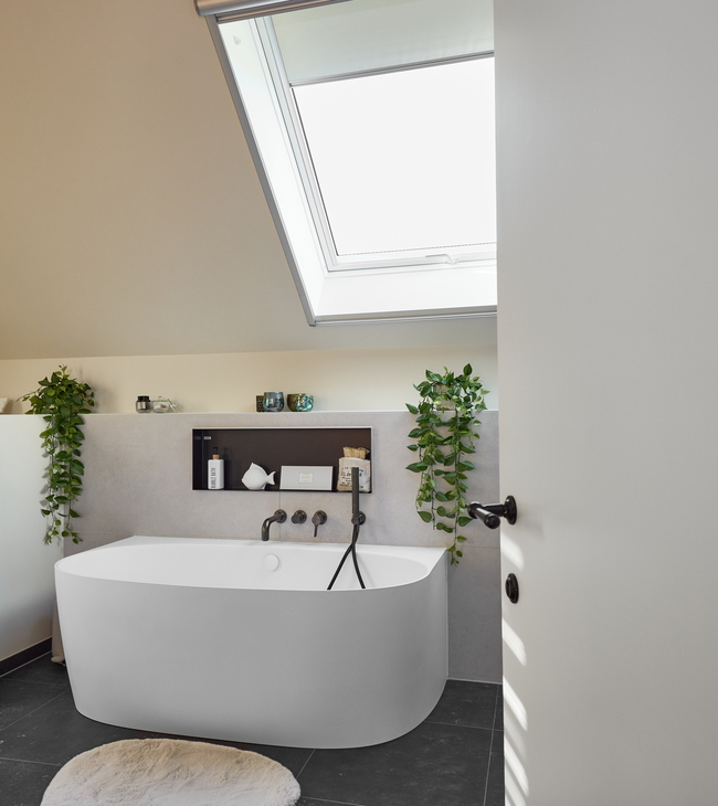 Rénovation de salle de bain moderne avec baignoire adossée au mur sous une fenêtre de toit, accents noirs et plantes vertes pour une ambiance fraîche.