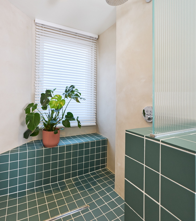 Douche avec carreaux verts et joint blanc, banquette intégrée, douche à effet pluie et plante près d’une fenêtre avec stores.