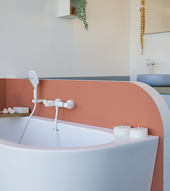 Baignoire d’angle îlot aux formes arrondies, avec robinetterie blanche et douchette, entourée d’accessoires chaleureux dans une salle de bain de style nordique.