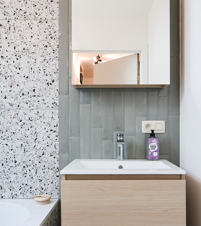 Meuble de salle de bain compact avec lavabo, armoire miroir et mur de carreaux verts allongés, à côté d’un habillage de baignoire en terrazzo.