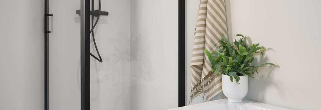 Douche moderne avec profils noirs à côté d’une baignoire blanche, aménagée avec des plantes et des accessoires pour un style de salle de bain épuré.